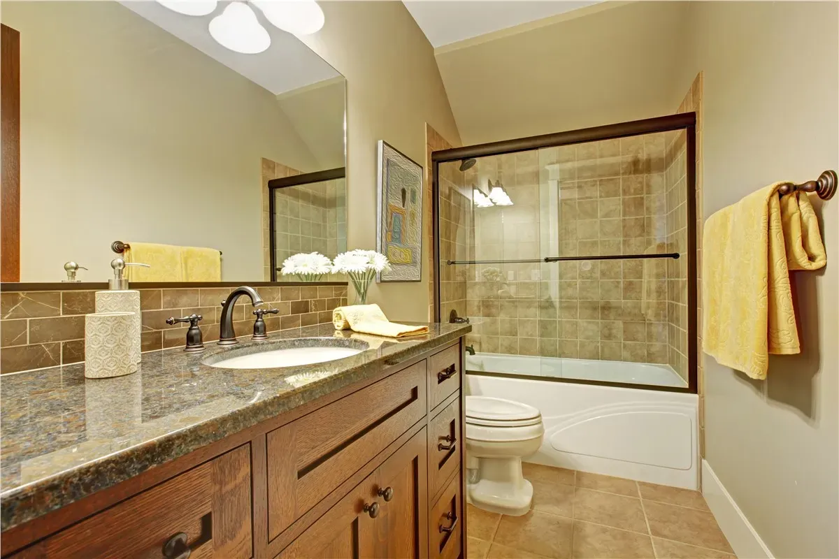 Warm-toned bathroom with wooden vanity, granite countertop, and vessel sink. Tiled shower with glass door, and yellow towels adding a cozy feel.
